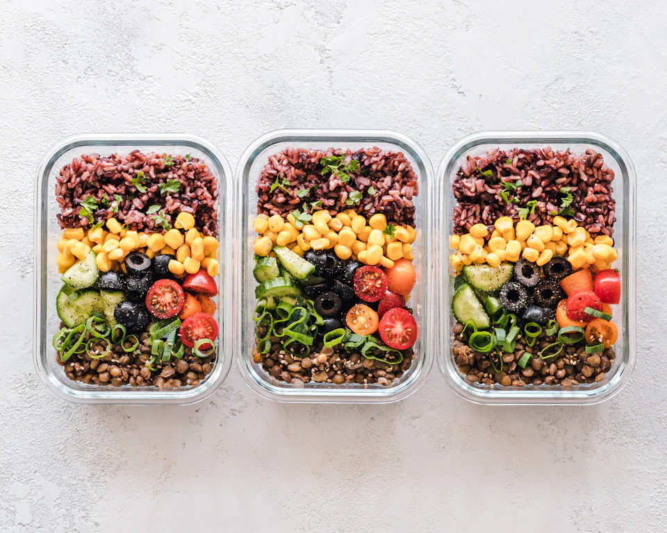 Three glass containers filled with rice, corn, olives, tomatoes, cucumbers, and lentils