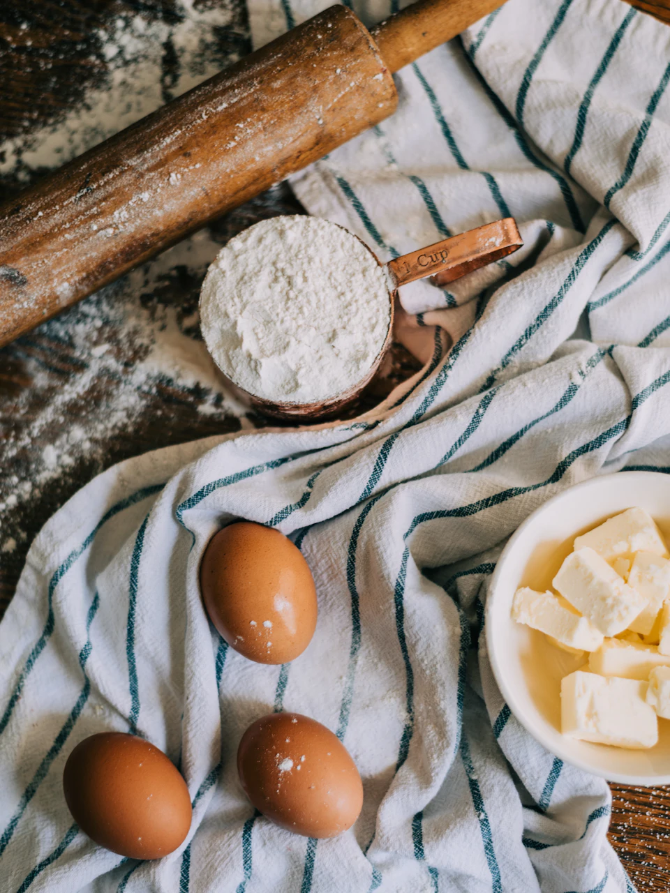 A measuring cup of flour, three brown eggs, and butter on a striped cloth