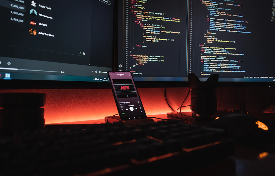 A smartphone displaying music on a desk with computer monitors showing code