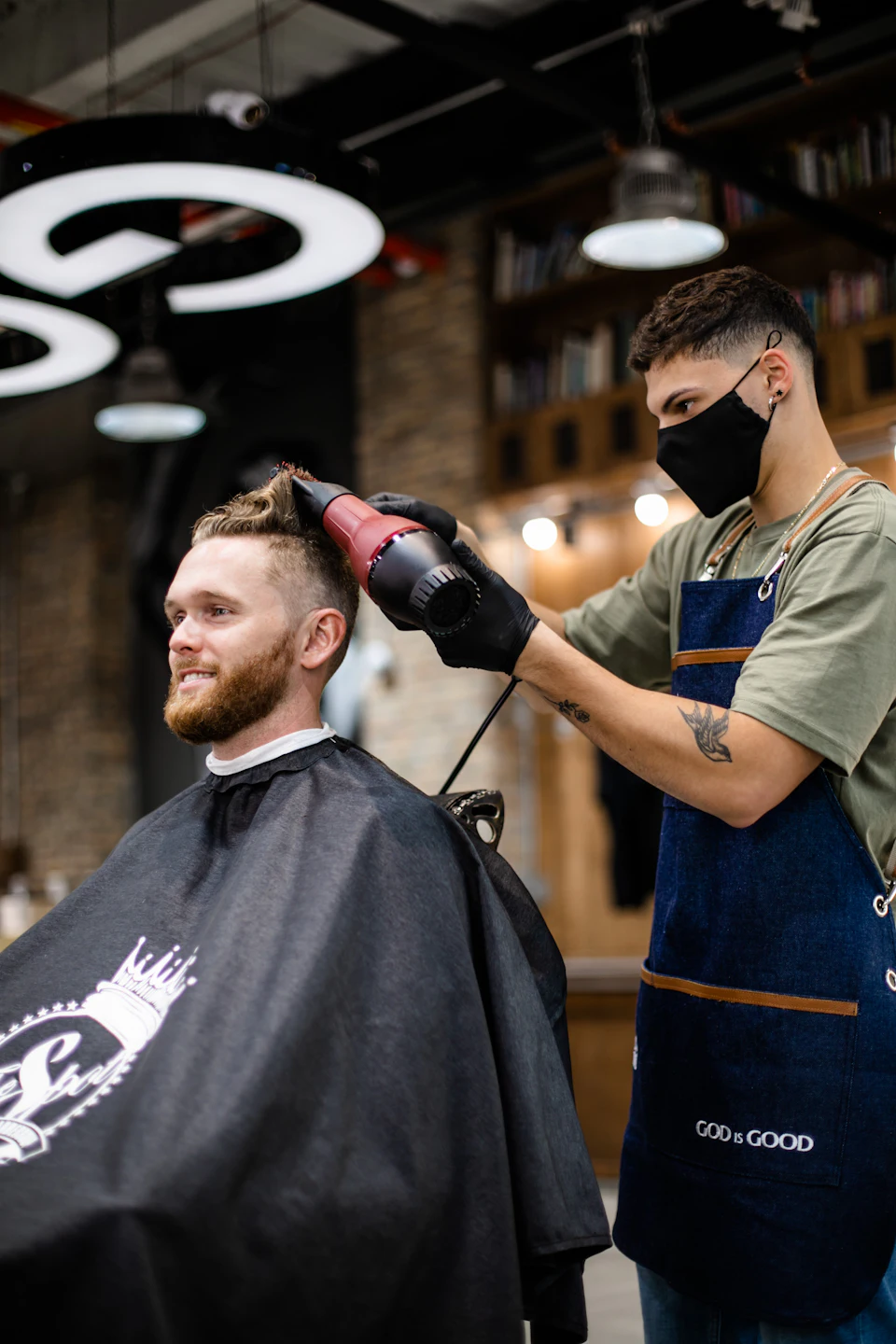 A barber in a denim apron blow-drying a man's hair in a salon