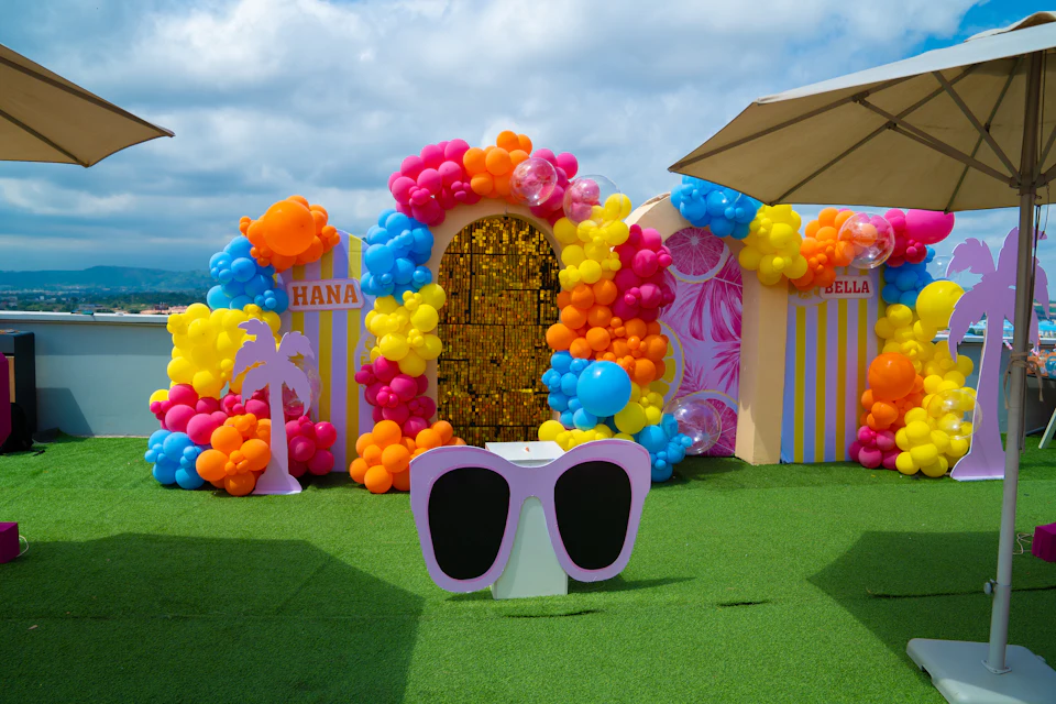 A colorful balloon arch and party backdrop on a green lawn under umbrellas